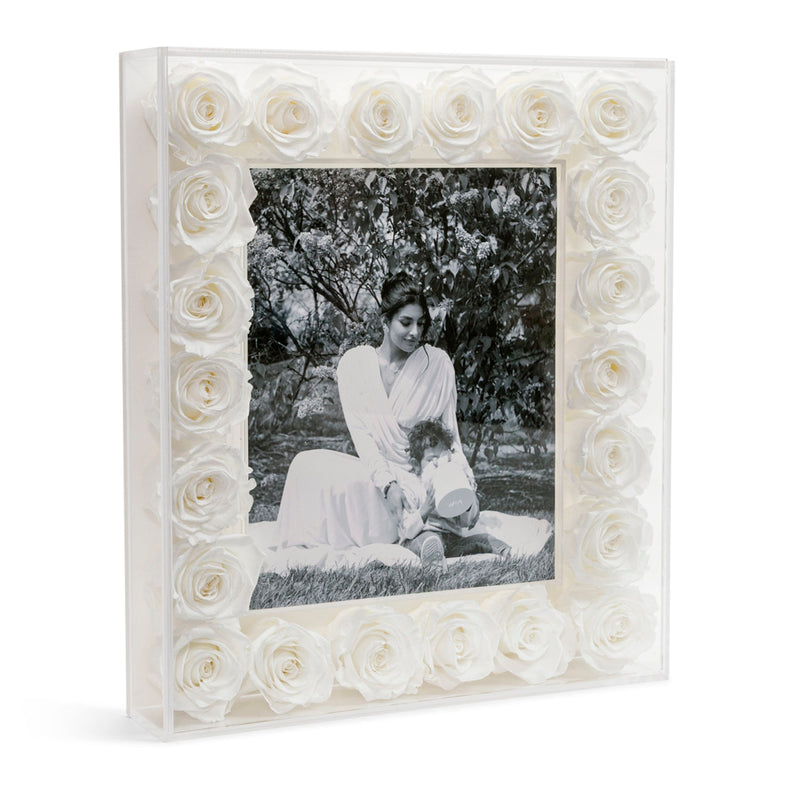 Photo frame with white roses surrounding a black-and-white photo of a woman sitting on grass with a child holding a container.